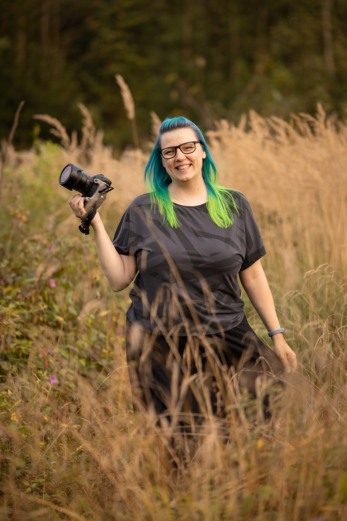 Die Fotografin Bettina Zajonc Farbhase Fotografie mit blau-grünen Haaren steht mit ihrer Kamera in der Natur und lächelt glücklich in die Kamera.